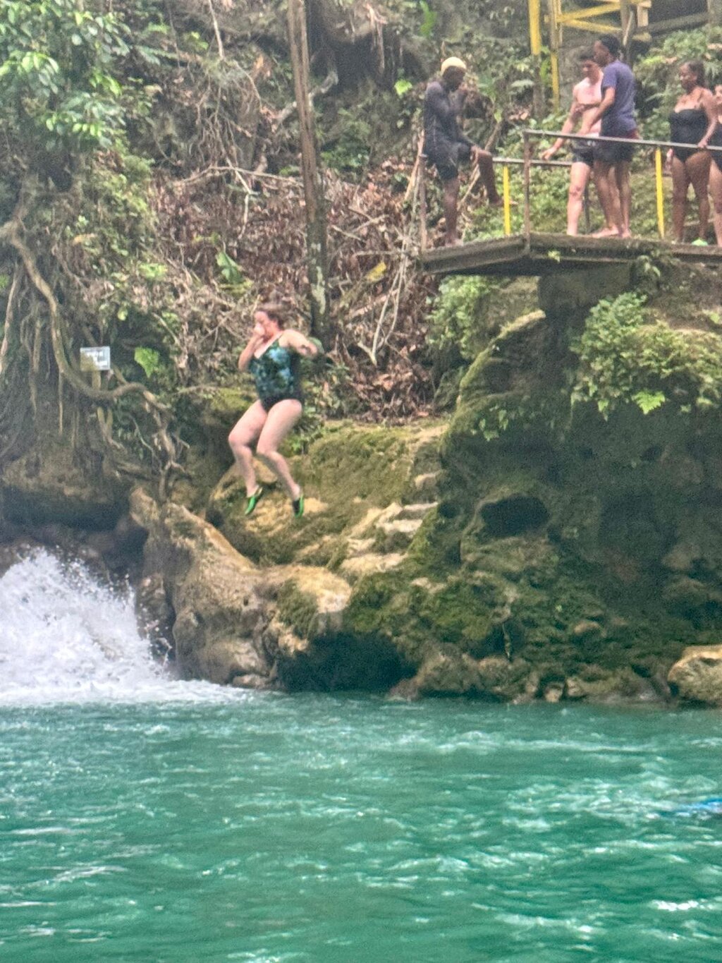 A tourist leaping off a cliff into the crystal-clear waters of Blue Hole Secret Falls, surrounded by lush greenery.